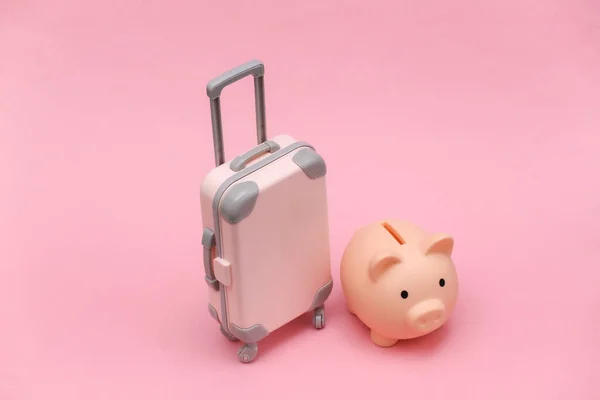 Travel or trip concept. Mini plastic travel suitcase and piggy bank on pink background. Minimal style