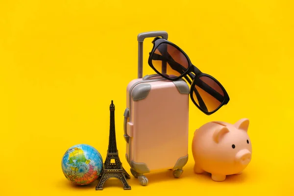 Traveled concept. Mini plastic travel suitcase with sunglasses and statuette of the Eiffel Tower, globe, piggy bank on yellow background.