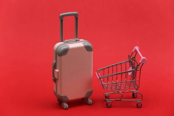 Travel and shopping concept. Mini plastic travel suitcase and supermarket trolley on red background.