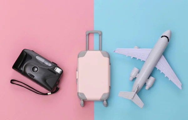 Travel flat lay. Mini plastic travel suitcase, camera and air plane on pink blue pastel background. Top view.