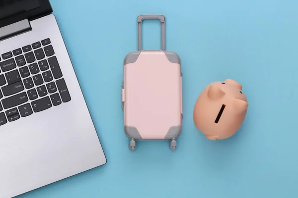 Flat lay vacation holiday and travel planing concept. Laptop and mini plastic travel suitcase, piggy bank on blue background. Top view
