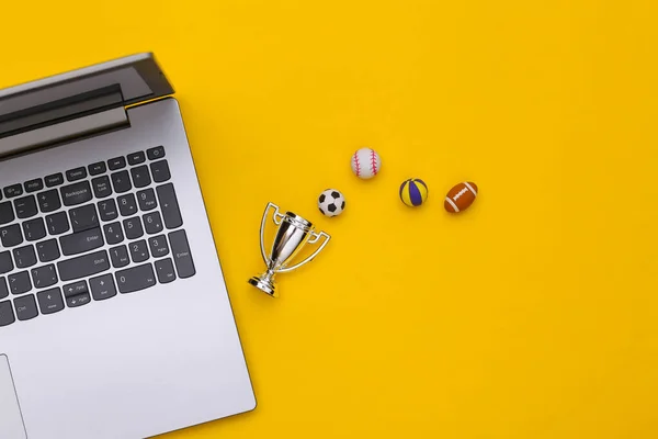 Online sports betting. Laptop, different sport ball and champion cup on yellow background with copy space. Top view. Flat lay