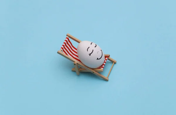 Chicken egg with hand drawn happy face on beach deck chair. Blue background. Summer rest, travel concept
