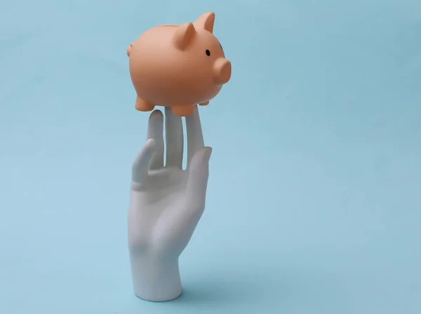 A white mannequin hand holds piggy bank on blue background. Finance, economy concept