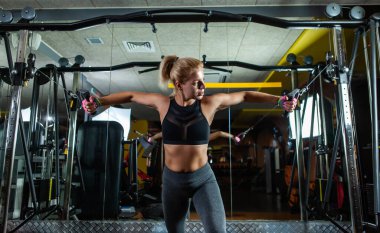 Zayıf fit sarışın kadın spor makinesinde kaslarını esnetiyor. Koyu renk spor salonunda egzersiz makinesi kablolu yayınıyla kız alıştırması yapıyor.
