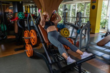 Genç ve çekici bir kadın spor salonunda egzersiz makinesinde çömelme egzersizi yapıyor.