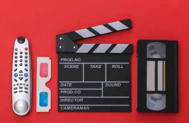 Film alkışlama tahtası, 3D gözlükler, video kaset ve kırmızı arkaplanda televizyon kumandası. Üst görünüm