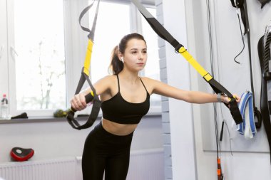 Genç, zinde bir kadın, ışık odasında kaslarını trx dirençli kemerlerle çalıştırıyor.