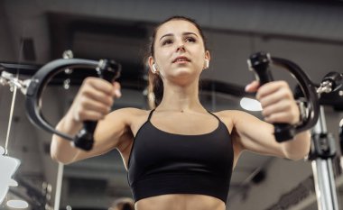 Genç, formda bir kadın, göğüs hatlarını egzersiz makinesinde çalıştırıyor. Kız modern spor salonunda çalışıyor.