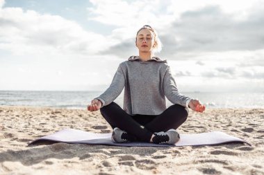 Spor giyimli çekici bir kadın güneşli bir günde kumsalda meditasyon yapıyor.