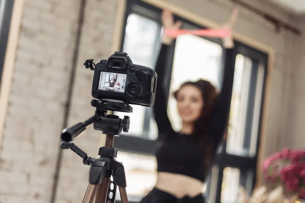 Young woman blogger with camera on tripod recording fitness blog and trains with fitness rubber bands. Fitness, sport and video blogging concept