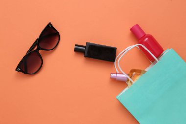 Güneş gözlüklü kağıt alışveriş çantası, turuncu pastel arka planda parfüm şişeleri. Üst görünüm