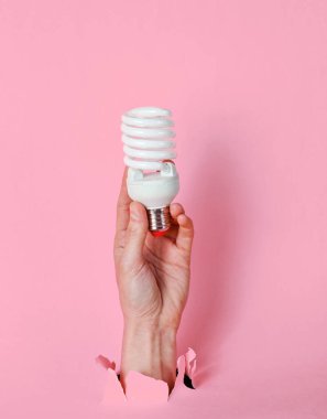 Kadın eli, yırtık pembe kağıdın içinde spiral ampulü tutuyor. Minimalist yaratıcı moda konsepti