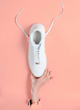 Kadın eli yırtık pembe kağıtların arasından beyaz spor ayakkabıları tutuyor. Minimalist yaratıcı moda konsepti