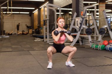Modern jimnastik salonunda elinde kettlebell olan kızıl saçlı, yoğun idman yapan bir kadın.