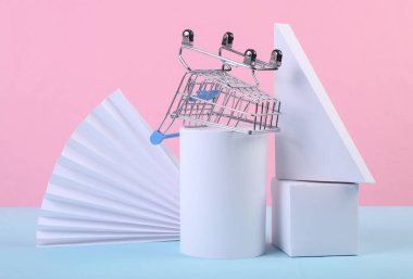 Alışveriş arabası ve geometrik şekiller mavi pembe pastel arka planda. Satış, ürün sunumu. Soyut arkaplan, kavram sanatı