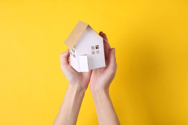 El, sarı arka planda küçük kağıttan ev heykelciği tutuyor. Koruma kavramı, güvenlik