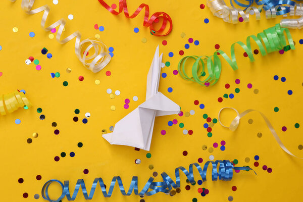 Origami rabbit with colored streamers and confetti on a yellow background. Easter celebration