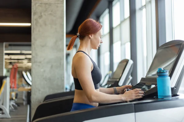 Portrait of a red-haired fitness woman in a modern gym. Fit slim woman works out on a treadmill, cardio workout, healthy lifestyle.