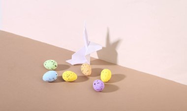 Yaratıcı Paskalya natürmortu. Origami tavşanı bej kahverengi arka planda renkli yumurtalar. Minimalizm. Moda gölgesi.