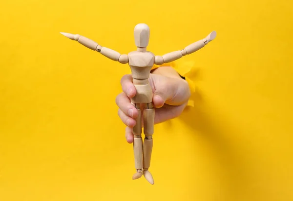 Female hand holds wooden puppet through torn hole yellow paper. Concept art. Minimalism