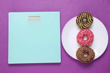 Weight loss concept. High-calorie donuts in plate and scales on purple background 