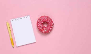 Weight loss plan, or recipe for cooking. Notepad with pen and donut on a pink background. Top view