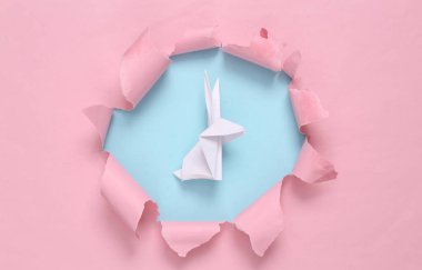 Origami rabbit through torn hole on blue-pink pastel background. Concept art. Pastel color trend. Minimalism