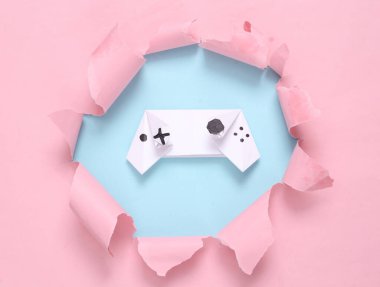 Origami gamepad through torn hole on blue-pink pastel background. Concept art. Pastel color trend. Minimalism