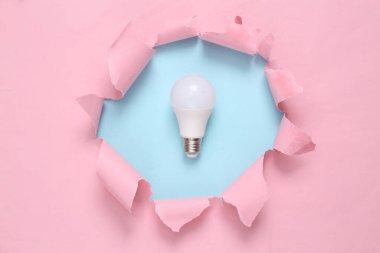 Led light bulb through torn hole on blue-pink pastel background. Business concept. Pastel color trend. Minimalism