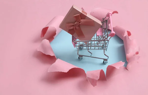 Shopping trolley with Gift box through a torn hole on blue-pink pastel background. Concept art. Pastel color trend. Minimalism