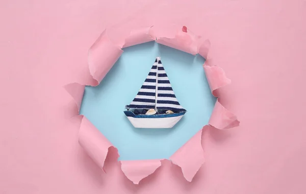 Toy Sailboat through a torn hole on blue-pink pastel background. Concept art. Pastel color trend. Minimalism