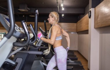 Spor kıyafetleri içinde uzun örgülü, zayıf, çekici bir kadın modern spor salonunda koşu bandında koşuyor.
