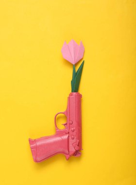 Sarı arka planda pembe bir silahın namlusundan çıkan Origami çiçeği. Yaratıcı romantik konsept. Minimalizm düzlük aşk. En küçük yerleşim. Üst görünüm