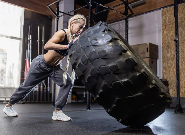 Fit female athlete flipping huge tire. Woman lifts a heavy wheel. Muscular young woman doing functional training exercise at gym.