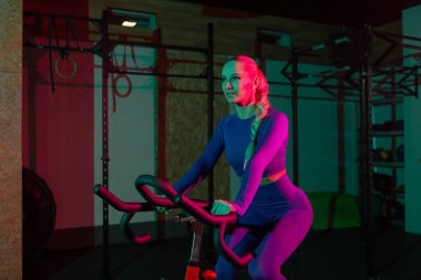 Güzel, genç, formda bir kadın spor salonunda staj bisikletinde pembe yeşil neon ışık gradyan ışıklarıyla kardiyo yapıyor. Spor, spor, yaşam tarzı konsepti