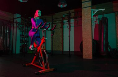 Güzel, genç, formda bir kadın spor salonunda staj bisikletinde pembe yeşil neon ışık gradyan ışıklarıyla kardiyo yapıyor. Spor, spor, yaşam tarzı konsepti