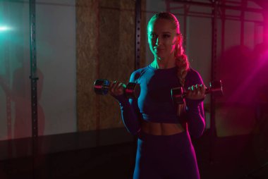 Genç, formda, zayıf bir kadın, elinde dambıllarla, spor salonunda, pembe, yeşil, neon renkli bir ışıkla kol kaslarını çalıştırıyor. Spor, spor, yaşam tarzı konsepti