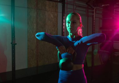 Genç, sıska kadın ellerinde kettlebell olan kasları jimnastik salonunda pembe yeşil ışıkla eğitiyor. Spor, spor, yaşam tarzı konsepti