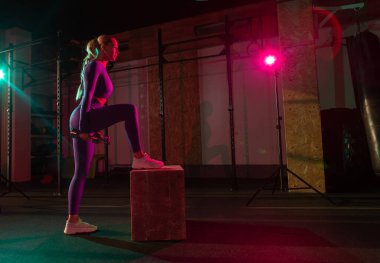 Kaslı kadın modern spor salonunda üzerinde pembe yeşil ışıklı dambıllar olan tahta bir kutuya basıyor. Spor, spor, yaşam tarzı konsepti. Fonksiyonel eğitim