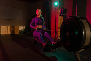 Kadın spor salonunda kürek makinesinde, pembe yeşil neon ışıkta kardiyo çalışması yapıyor. Sportif kız fitness kulübünde çalışıyor. Sağlıklı yaşam tarzı