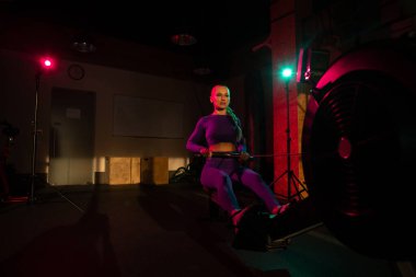 Kadın spor salonunda kürek makinesinde, pembe yeşil neon ışıkta kardiyo çalışması yapıyor. Sportif kız fitness kulübünde çalışıyor. Sağlıklı yaşam tarzı
