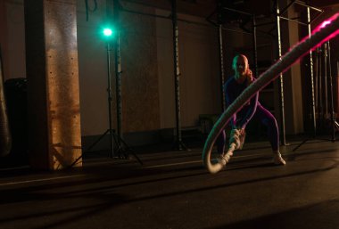 Atletik güçlü kadın modern spor salonunda savaş halatıyla egzersiz yapıyor. Pembe yeşil neon ışık. Fonksiyonel eğitim