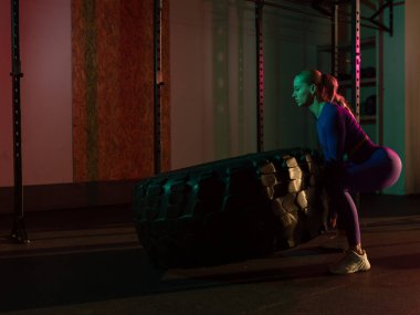 Fit bir bayan sporcunun lastiği patinaj çekiyor. Pembe neon ışıkta ağır bir tekerleği kaldıran bir kadın. Spor salonunda egzersiz yapan kaslı genç bir kadın..