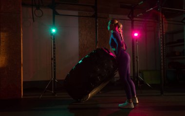 Fit bir bayan sporcunun lastiği patinaj çekiyor. Pembe neon ışıkta ağır bir tekerleği kaldıran bir kadın. Spor salonunda egzersiz yapan kaslı genç bir kadın..