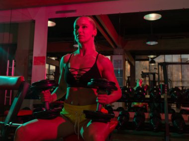 Spor giyim sektöründe formda bir kadın pembe yeşil ışıklı bankta otururken ağır halterlerle antrenman yapıyor. 