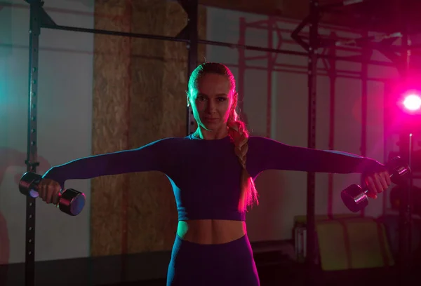 体形苗条的年轻女子手握哑铃 在健身房的粉色绿色霓虹灯下锻炼肩部肌肉 生活方式概念 — 图库照片