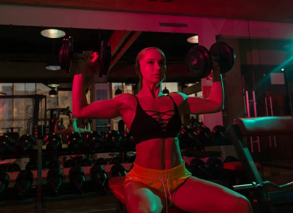 Spor giyim sektöründe formda bir kadın pembe yeşil ışıklı bankta otururken ağır halterlerle antrenman yapıyor. 