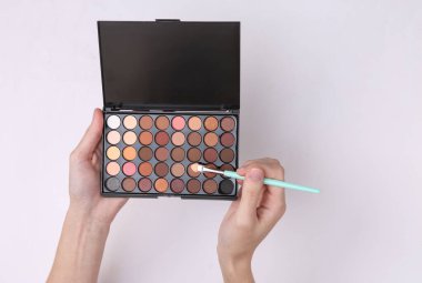 Göz farı paleti tutan kadın elleri ve beyaz arka planda makyaj fırçaları.