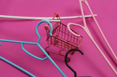 Pembe arka planda mini alışveriş arabası olan plastik askılar. Moda düzeni.